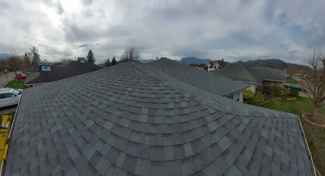 Aerial drone view of a completed residential roof installation with gray architectural asphalt shingles, featuring Cascade mountain backdrop and evergreen forest typical of Chilliwack area.