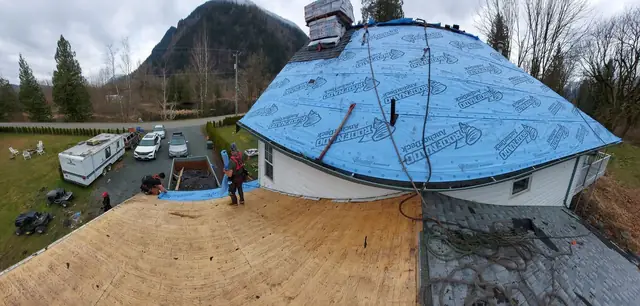 Aerial view of residential roof decking installation with full crew visible, blue underlayment applied, mountain backdrop in alpine Fraser Valley setting.