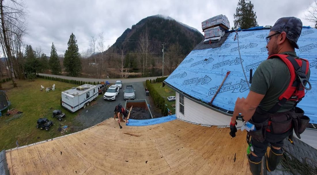 Johnny Peters of Fraser Roofing standing on a roof during a full roof replacement on Seabird Island BC, with Roofnado ArcherDeck underlayment installed and shingle bundles staged at the ridge, Fraser Valley mountains in the background