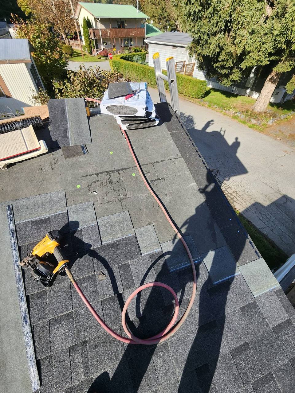 Mid-installation on a Fraser Valley residential re-roof, a pneumatic nailer and fresh shingle bundles at the ready as the Fraser Roofing crew works toward the ridge cap on a clear autumn day.