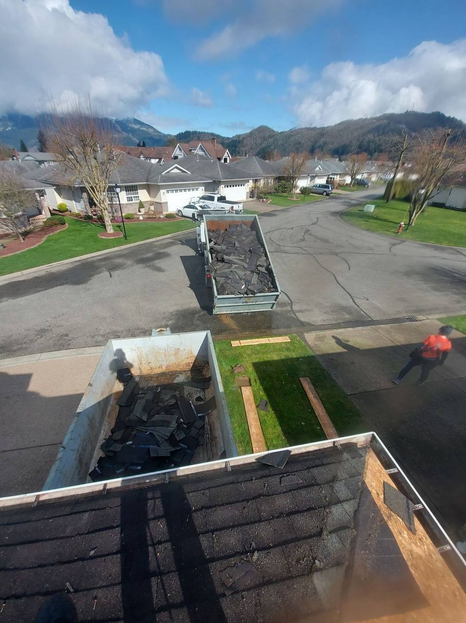 Fraser Roofing crew performing asphalt shingle tear-off on a residential home in Agassiz BC, with debris bins loaded with old roofing material and mountain backdrop of the Fraser Valley