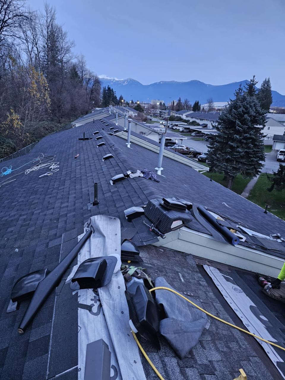 Dusk rooftop view of a near-complete asphalt shingle reroof on a multi-unit residential complex in Chilliwack BC with ridge vents installed and snow-capped mountains in background