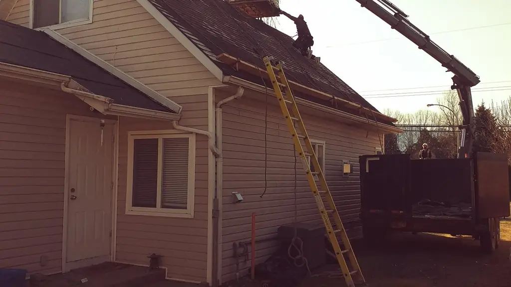 A knuckle-boom crane truck lifts a bundle of shingles up to two roofers working the steep gable roof of a mauve-sided house at golden hour. Fraser Roofing