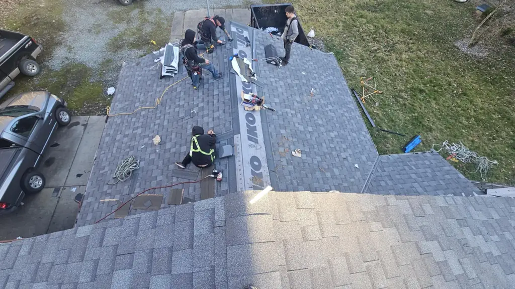 Overhead view down onto a driveway and roof where four crew members work at a hip roof section, one sitting cross-legged with a cordless drill. Fraser Roofing