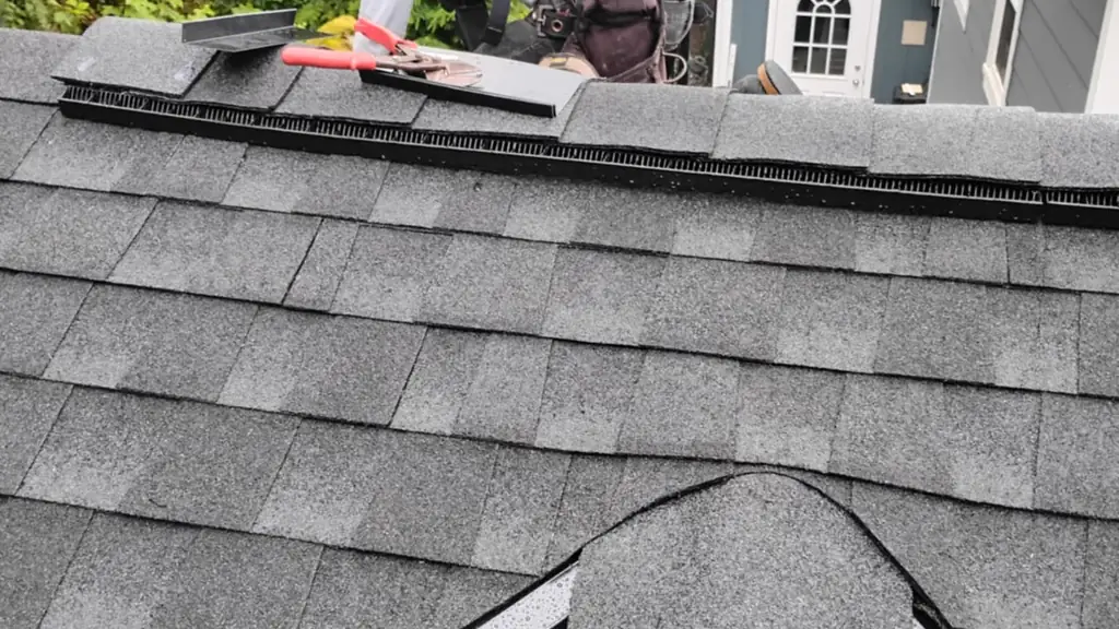 A roofer wearing a safety harness is crouched near the ridge of a gray dimensional shingle roof. Fraser Roofing
