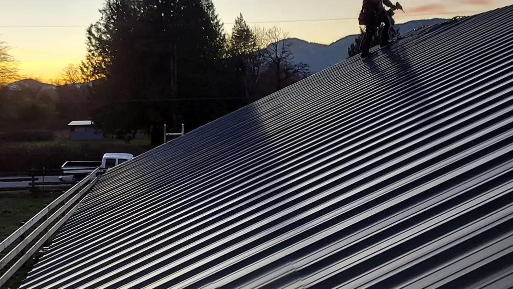 A worker is silhouetted near the ridge of a dark grey standing-seam metal roof at dusk, holding what looks like a cordless drill or impact driver. Fraser Roofing