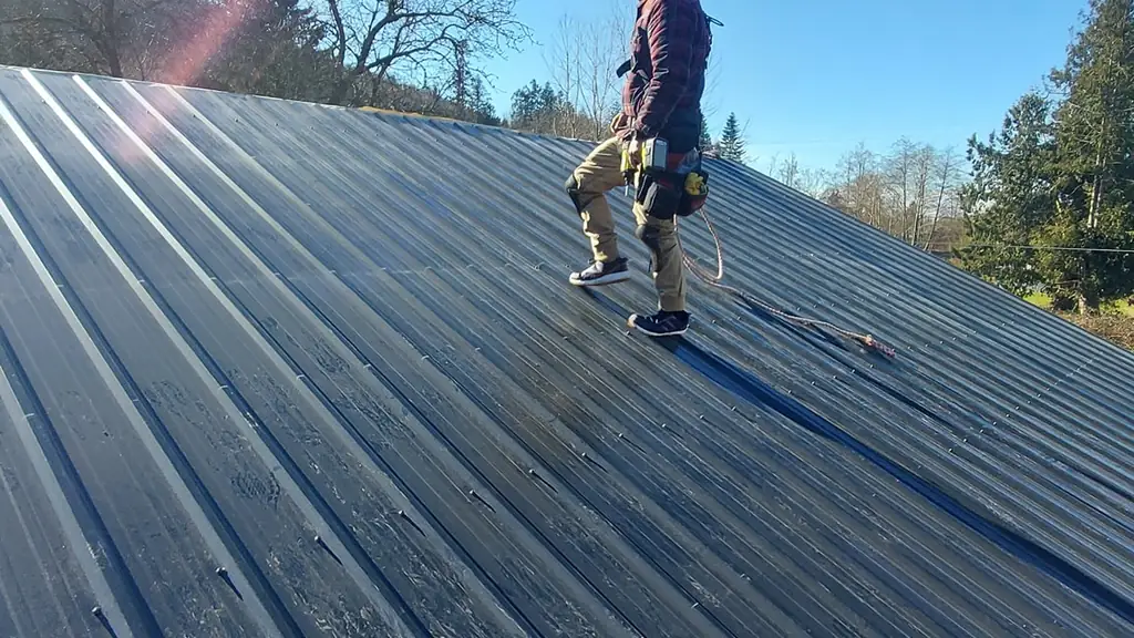 A worker in a plaid shirt and tool belt stands on a corrugated metal roof with a safety rope trailing behind him. Fraser Roofing