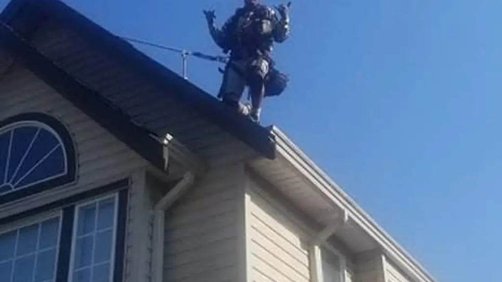Looking up from the ground at a worker standing on a roof ridge in a full-body safety harness, both arms raised with a thumbs up. Fraser Roofing