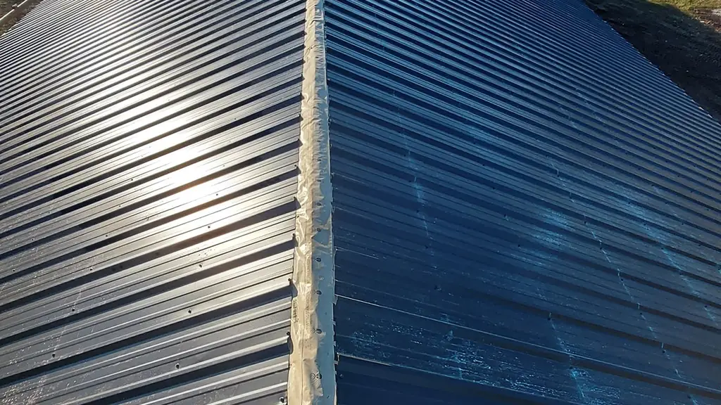 Standing on the ridge of a long, completed dark standing-seam metal barn roof, with a fall-arrest anchor and rope visible in the foreground. Fraser Roofing