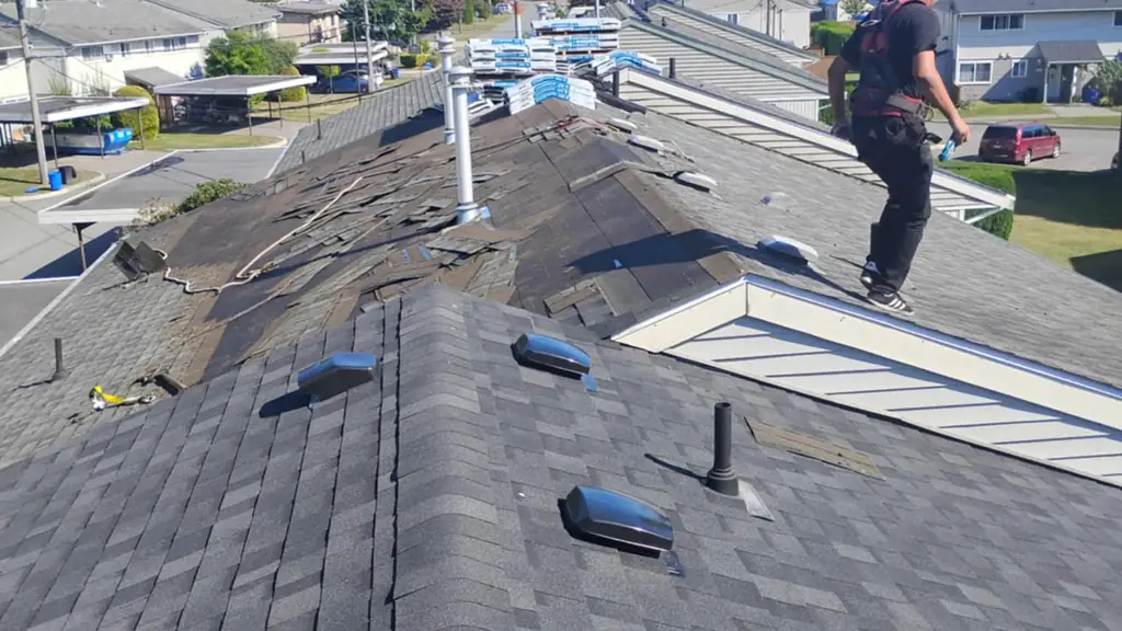 A crew member in a ball cap and safety harness walks along the ridge of a finished dark grey shingled roof under a bright blue sky with distant. Fraser Roofing