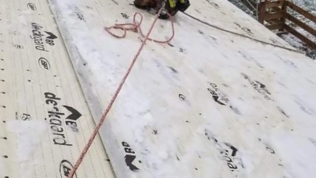 A smiling worker kneels on a snow-dusted roof covered in synthetic underlayment printed with a brand name, gripping a safety rope. Fraser Roofing