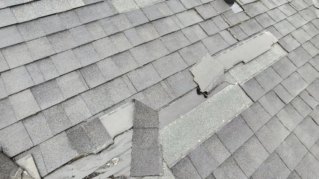 Wind-lifted and torn asphalt shingles on a roof slope beside a plumbing vent. Fraser Roofing