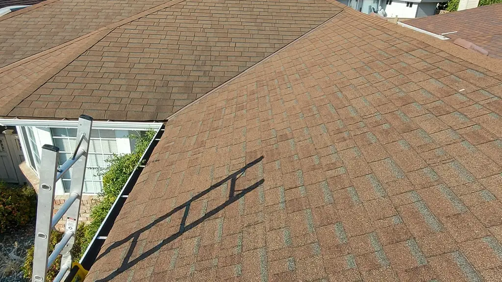 A view along a brown asphalt shingle roof ridge with a ladder and its long shadow crossing the slope. Fraser Roofing