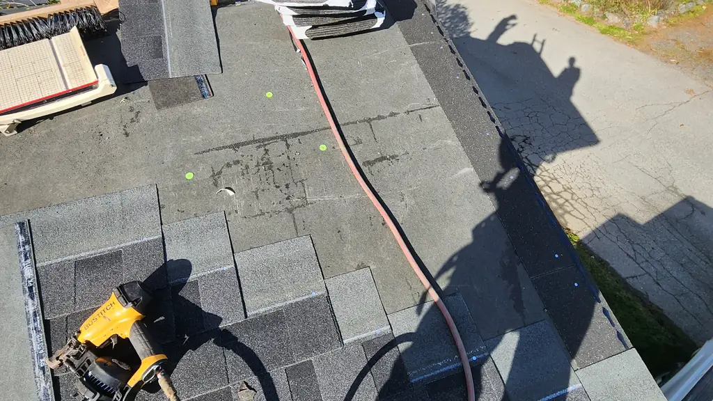 A near-identical companion shot to the previous one: a yellow Bostitch pneumatic nail gun with a coiled pink air hose resting on new grey shingles. Fraser Roofing
