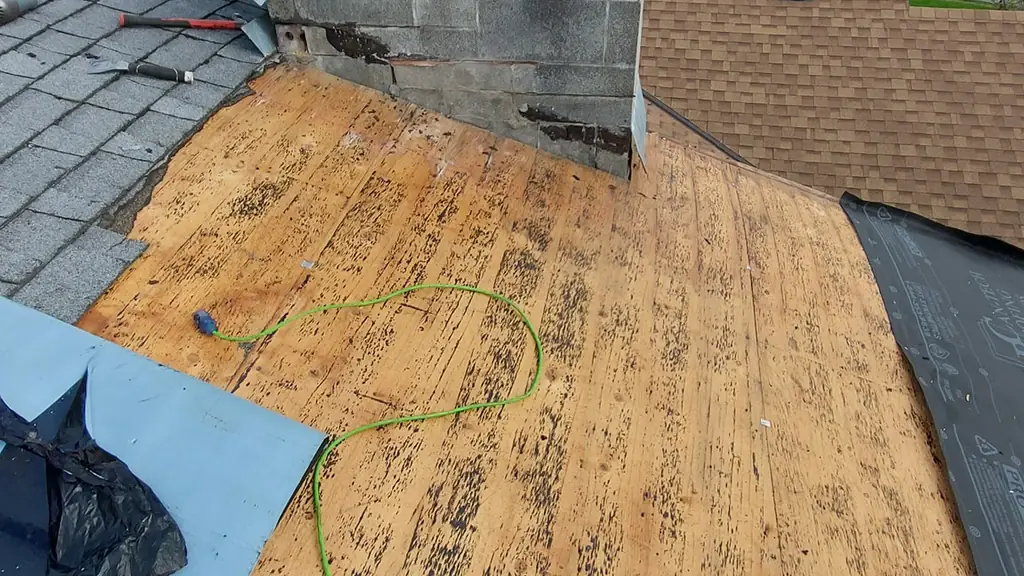 A large concrete-block chimney with an open spark-arrestor cap and two round vent caps rises from a roof stripped down to plywood decking. Fraser Roofing
