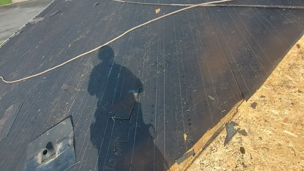 Aerial view down an old black tar-paper roof with a worker's own shadow cast large across the surface, and a second. Fraser Roofing