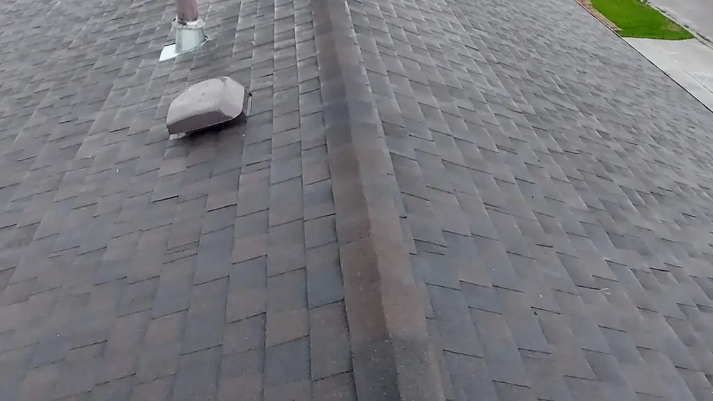 An on-roof view along the ridge cap of a brown, aged asphalt shingle roof, with vents and a stone chimney visible along the peak. Fraser Roofing