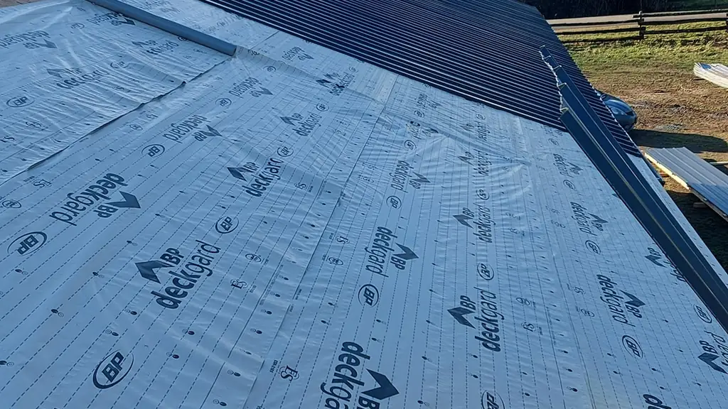 Standing on a partially installed dark standing-seam metal roof, with white BP Deckgard synthetic underlayment covering the rest of the deck. Fraser Roofing
