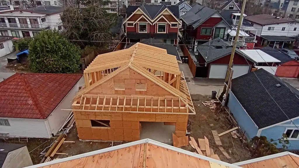 Aerial view taken from atop a freshly-decked plywood roof, looking down over a new-construction house whose roof is at the framed-rafters stage (no. Fraser Roofing