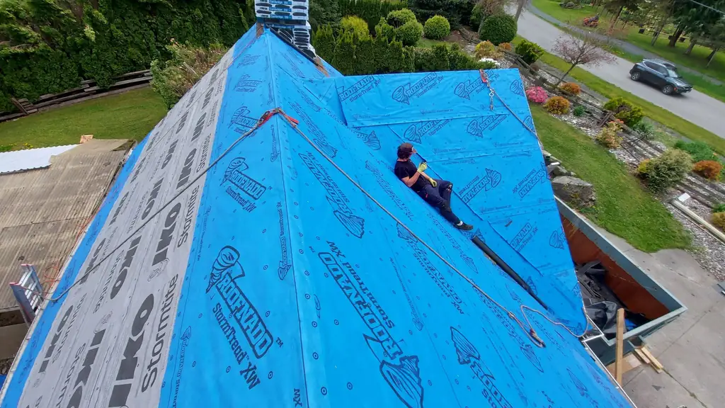 A worker reclines on a steep roof slope covered in blue 'Roofnado StormTread' synthetic underlayment, clipped into a rope for fall protection. Fraser Roofing
