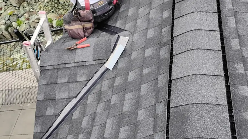 Close-up of a worker in a white t-shirt and red safety harness, kneeling on a roof edge backed by a mossy rock outcrop and ferns. Fraser Roofing