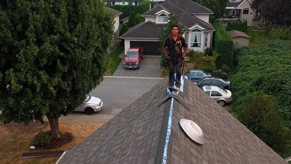 A worker stands confidently on a roof ridge with hands on hips, wearing a full harness over a dark shirt, looking directly at the camera. Fraser Roofing