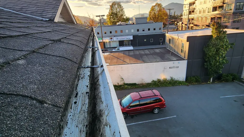 Looking down along a stained white fascia board and gutter line at the edge of an asphalt shingle roof toward a lit 'Downtown Business Centre'. Fraser Roofing