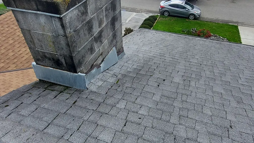 An on-roof shot of a large concrete-block chimney topped with a mossy cap, next to a grey shingle roof. Fraser Roofing