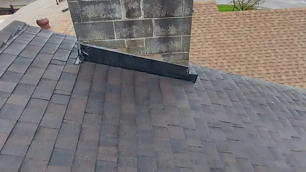 A large mossy concrete-block chimney with an open-top spark-arrestor cap rises above a tan asphalt shingle roof. Fraser Roofing