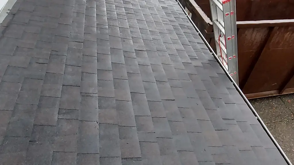 A view looking along the eave of a dark, worn asphalt shingle roof toward a wooden dump trailer with an aluminum ladder leaning into it. Fraser Roofing