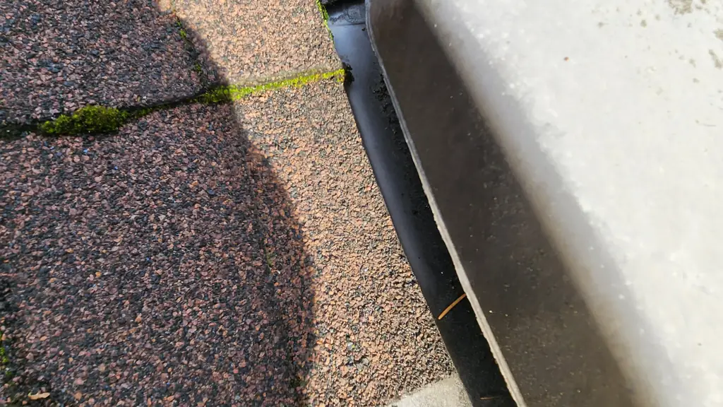 An extreme close-up shows a white plastic gutter downspout outlet at a roof corner, with moss growing in the shingle grooves nearby. Fraser Roofing