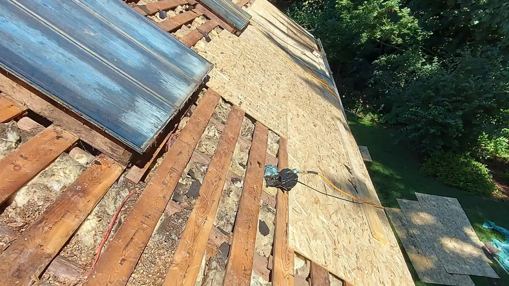 A steep roof mid tear-off shows exposed rafters with pink fiberglass insulation between them. Fraser Roofing
