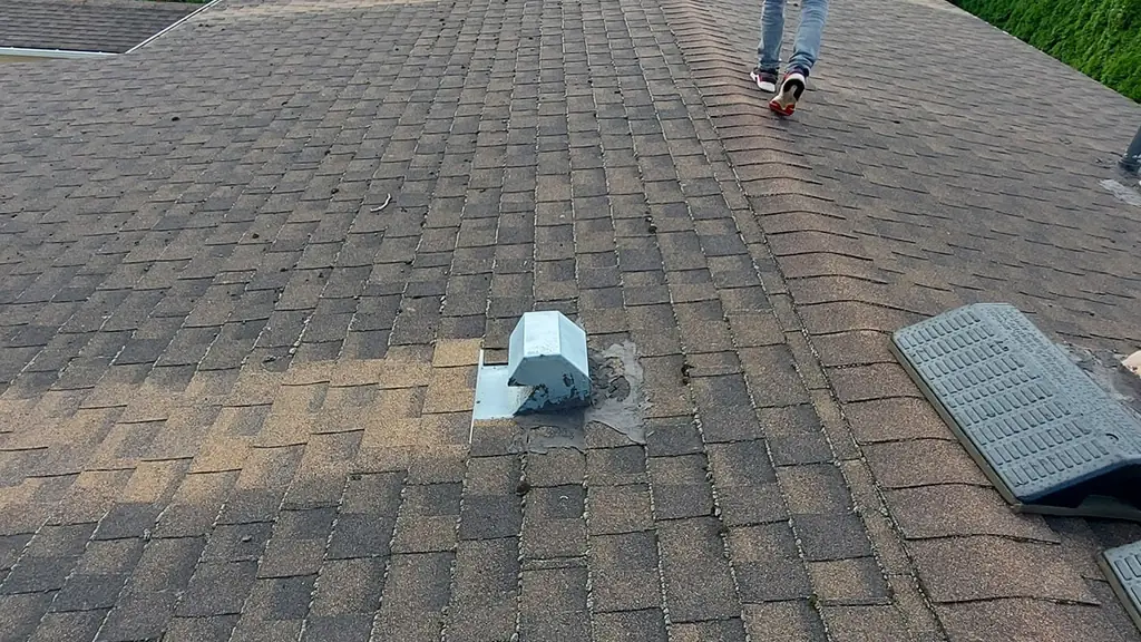 A worker in a red, white and black hoodie walks away from the camera along a worn, aged asphalt shingle roof toward a pipe boot vent. Fraser Roofing