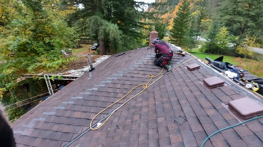 A worker wearing a tool belt kneels on a brown asphalt shingle roof next to a brick chimney, working on something at roof level. Fraser Roofing