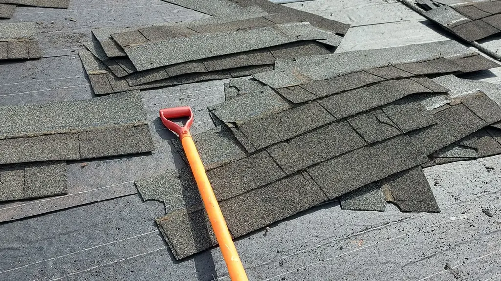 A view up a roof slope covered in torn, curling grey shingles being removed, exposing patches of plywood decking and felt underlayment near the ridge. Fraser Roofing