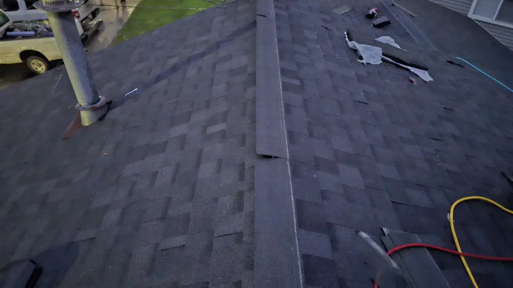 A dim, dusk-lit shot looking down a dark grey asphalt shingle roof ridge, with vehicle work-lights glowing on a wet street below. Fraser Roofing