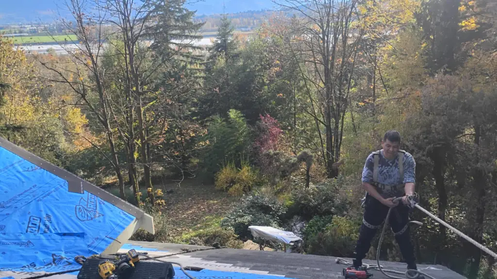 A worker in a harness stands on a roof, smiling at the camera while pulling a thick rope over his shoulder. Fraser Roofing