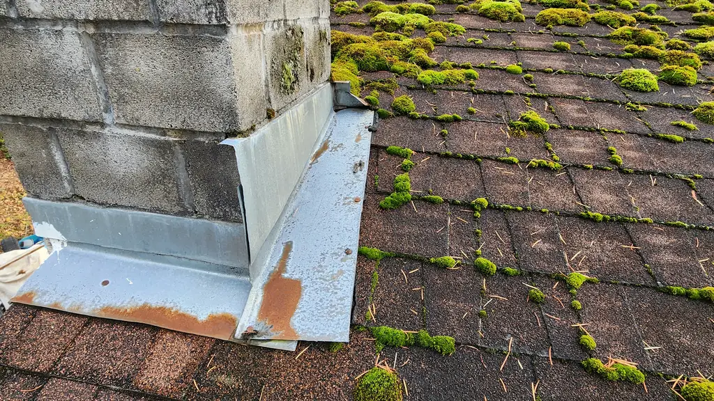 A close view of a concrete-block chimney base where new galvanized metal flashing (some sections rust-stained) meets an asphalt shingle roof. Fraser Roofing