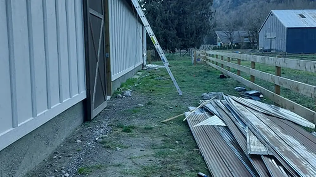 Ground-level view alongside a light gray metal-sided barn, with an extension ladder leaning against the roofline. Fraser Roofing