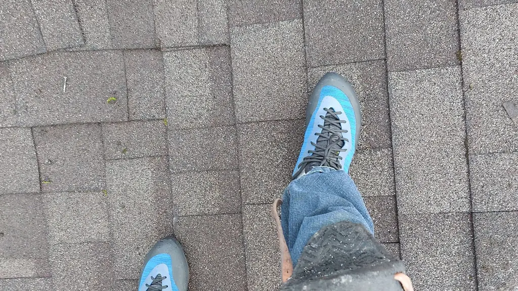 A first-person shot looking straight down at the photographer's own feet, wearing blue-and-black climbing-style roofing shoes and rolled jeans. Fraser Roofing
