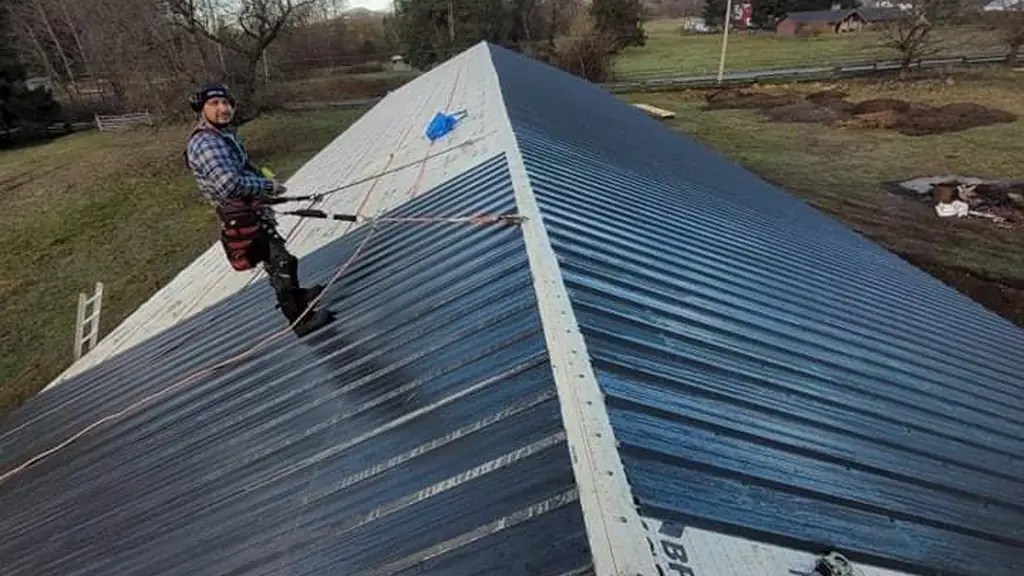 A worker wearing a plaid shirt, tool belt and safety harness stands near the ridge of a dark grey corrugated metal roof. Fraser Roofing