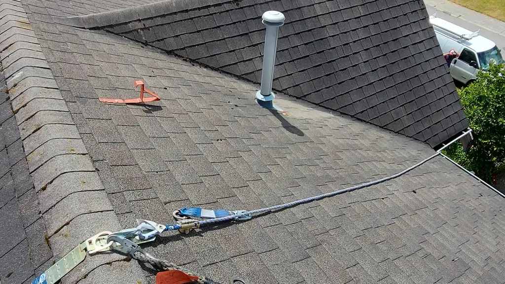 A safety rope lifeline runs along a roof ridge, threaded through carabiners and rope grabs, passing near a plumbing vent stack. Fraser Roofing
