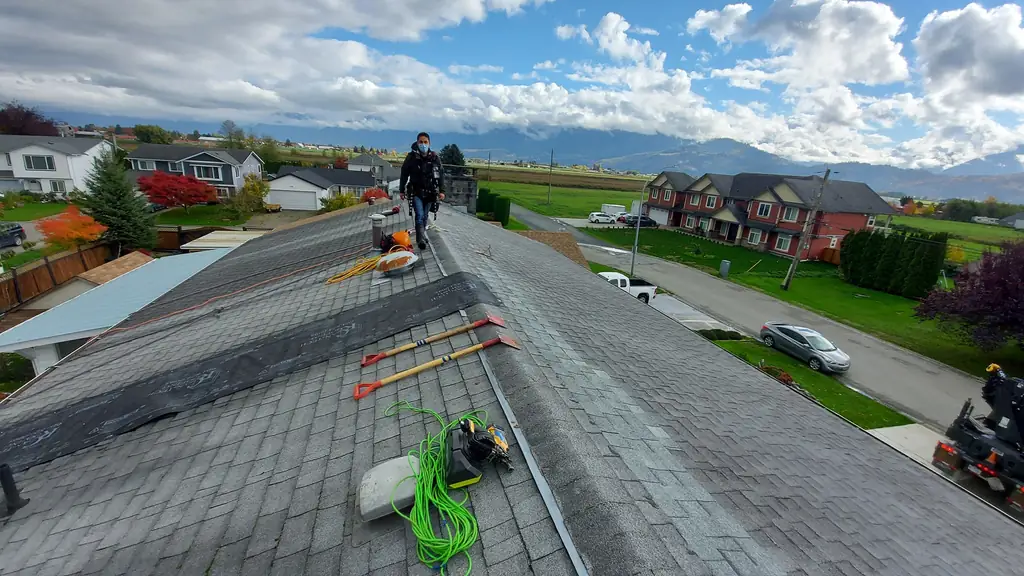 A wide shot along a roof ridge under a partly cloudy sky, with a worker wearing a safety harness and a face mask walking toward the camera near. Fraser Roofing