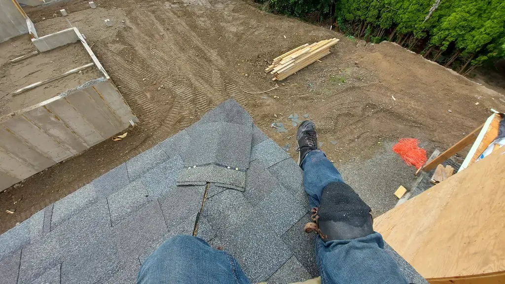 A point-of-view shot from the edge of a roof on a new-construction site, looking down past a worker's own legs and boots to a bare dirt lot below. Fraser Roofing