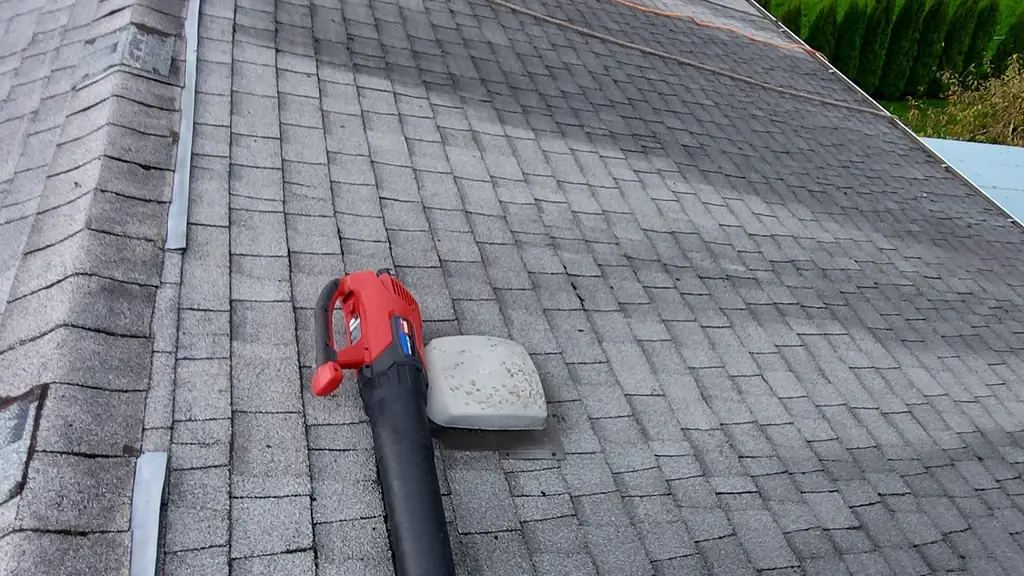A worker in a harness stands on a roof ridge far in the distance, while a leaf blower and a vent boot sit in the foreground on a grey shingle roof. Fraser Roofing