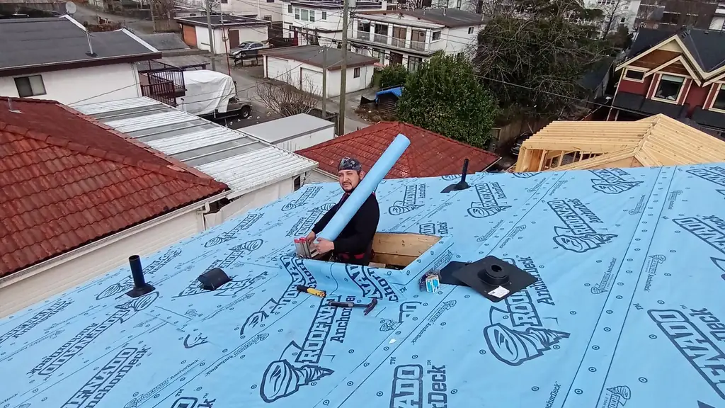 A worker leans out of a roof hatch/chimney chase cut into blue 'Roofnado AnchorDeck' synthetic underlayment. Fraser Roofing