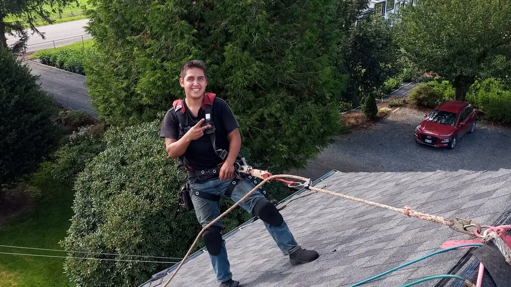 A young man in a black t-shirt and harness stands on an asphalt shingle roof ridge, smiling at the camera and making a peace sign. Fraser Roofing