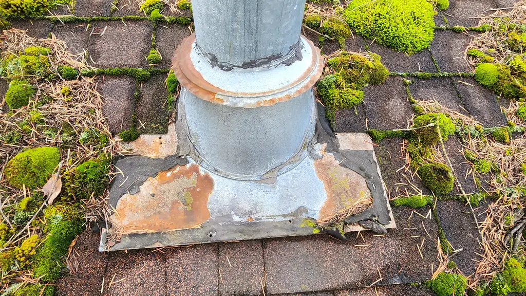 An extreme close-up of an old galvanized pipe vent base with a rusted metal collar, surrounded by thick. Fraser Roofing