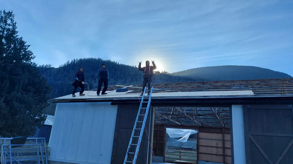 Three workers stand on the open roof deck of a barn or outbuilding under construction, with exposed rafters and plywood sheathing visible around them. Fraser Roofing