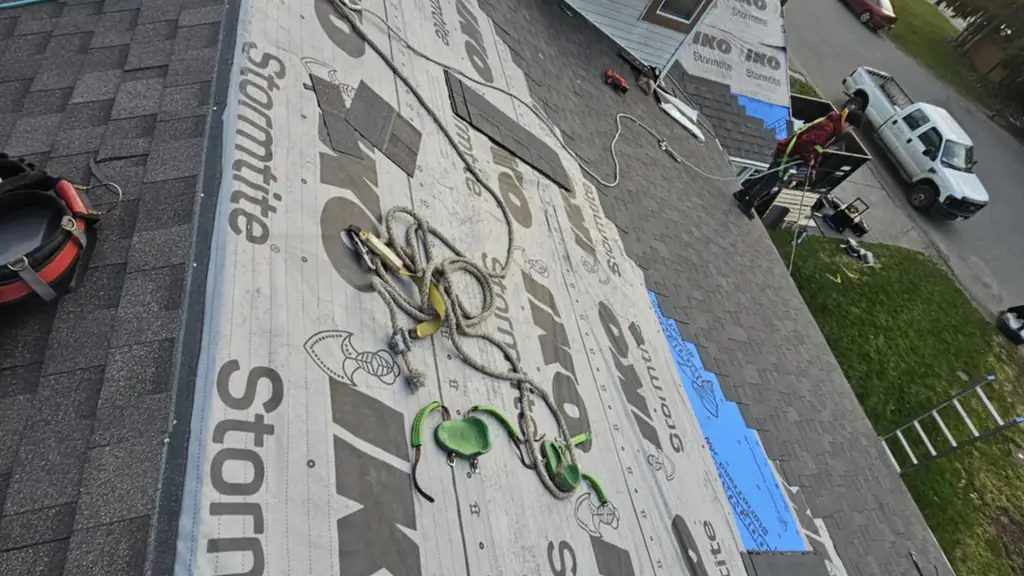 Looking down a roof ridge covered in IKO Stormtite underlayment with a rope-and-pulley rigged to a safety anchor, tools scattered nearby. Fraser Roofing